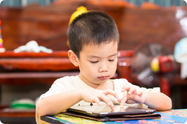 Young Asian boy sitting at desk playing on and ipad.