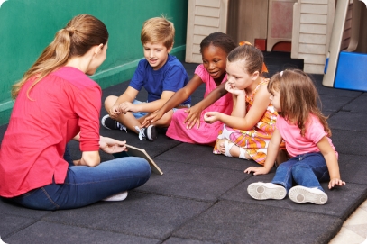 Toddlers sitting on a mat during story time or circle time looking intently at a story book being held by a teacher.