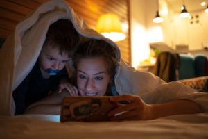 Young mother with young child under a blanket, reading from a smart device.